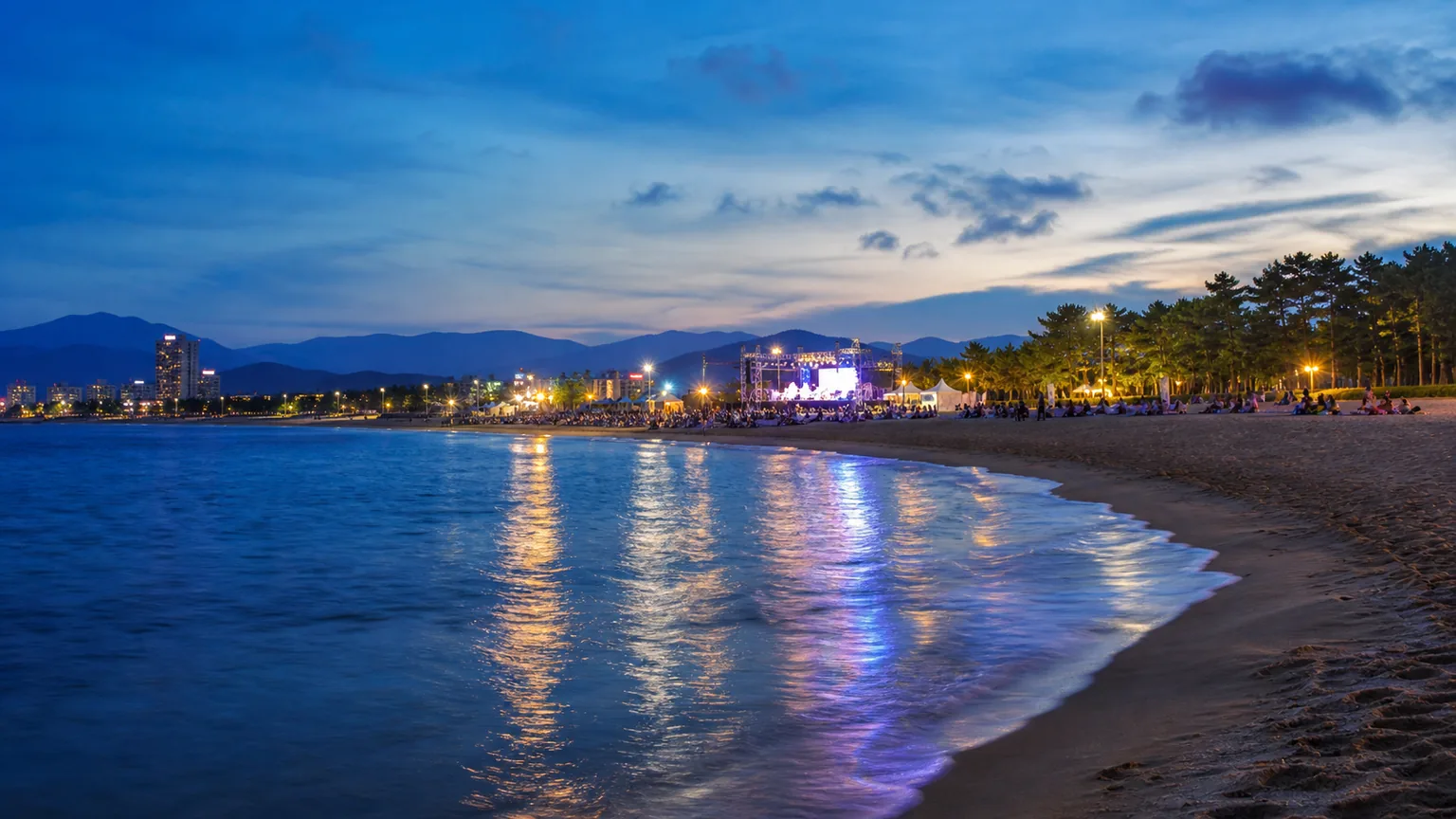 Editorial night view of a Korean East Coast beach with a distant festival stage and shoreline lights.