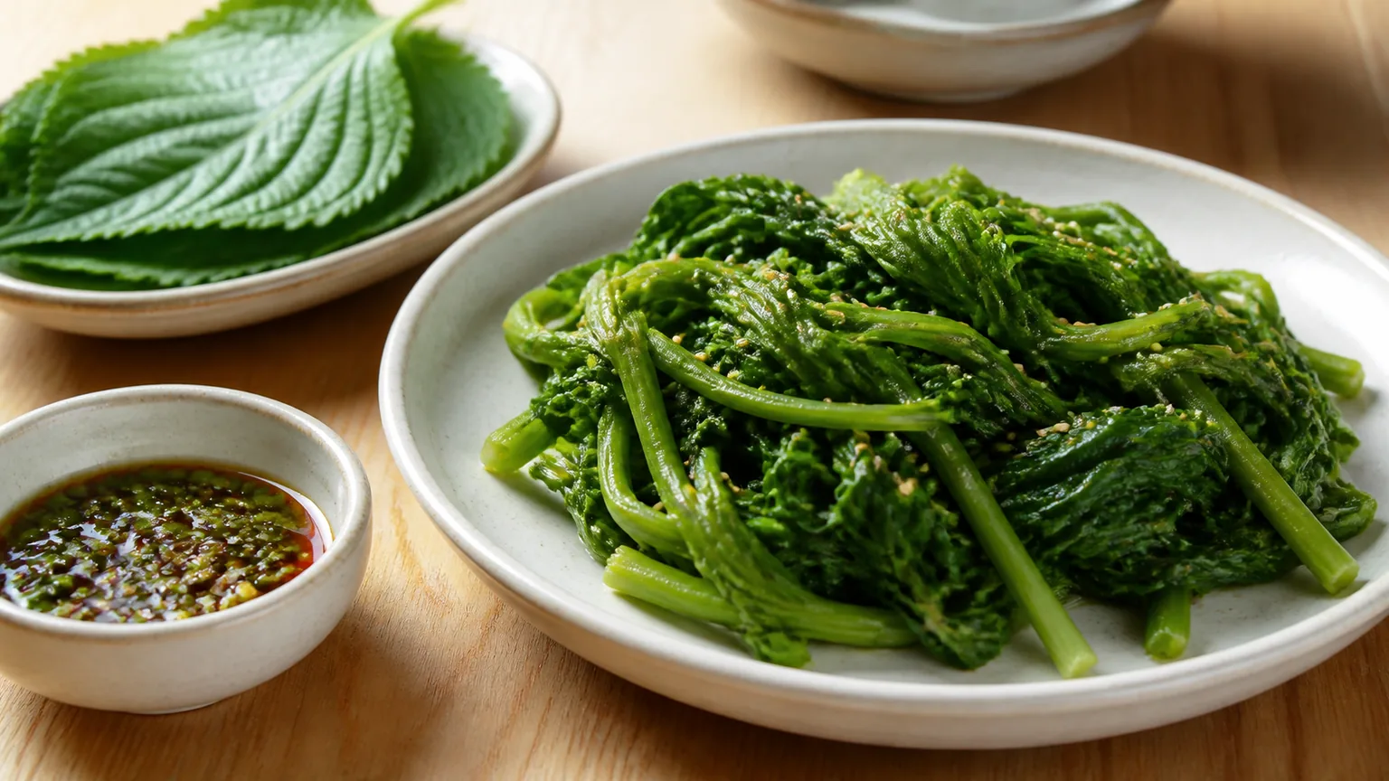 Seasoned spring greens beside perilla leaves and soy-based dipping sauce on a Korean table